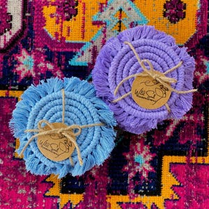 May include: Two handmade macrame coasters, one light blue and one purple, are displayed on a colorful patterned rug. Each coaster is round with a fringed edge and tied with twine and a small tag. The tags have the text "Wild Soul Creations by Mimi."