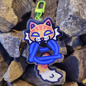 Può includere: Un portachiavi a forma di gatto colorato con un design a triangolo blu. Il gatto ha una pelliccia arancione e rosa con dettagli blu e gialli. Ha un fermaglio verde per attaccarlo a chiavi o borse. Lo sfondo è un insieme di rocce grigie.