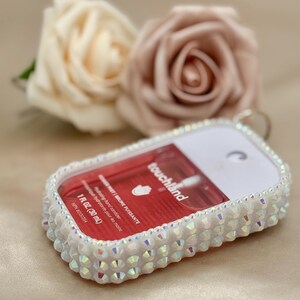May include: A white hand sanitiser spray bottle with iridescent rhinestones around the edges. The bottle contains a red liquid and has the text "touchland" and "1 FL OZ. (30 mL)". Two artificial roses are in the background.