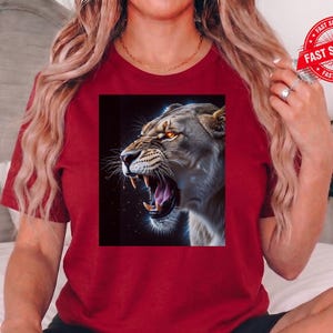 May include: A maroon t-shirt featuring a detailed graphic of a roaring lioness with glowing orange eyes. The lioness is set against a dark background, with a focus on its open mouth and sharp teeth. The shirt is a solid color.