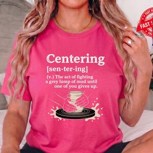 May include: A hot pink t-shirt with the word "Centering" in white, along with a definition and illustration of hands shaping clay on a pottery wheel. The shirt also has a "FAST SHIPPING" sticker.