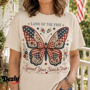 May include: A cream-coloured t-shirt featuring a large butterfly graphic with red, white, and blue patterns, including stars, stripes, and gingham. The text reads LAND OF THE FREE above the butterfly and Spread Your Stars & Stripes below it.
