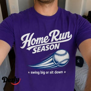 May include: A purple short-sleeve t-shirt featuring the white text Home Run SEASON above a graphic of a baseball in motion with blue motion lines. Below the baseball, the text reads swing big or sit down between two white stars.