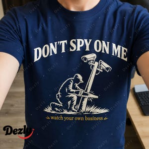 Może przedstawiać: Granatowa koszulka z białym napisem DON'T SPY ON ME u góry. Poniżej grafika przedstawia klęczącą osobę piłującą słup z dwiema kamerami bezpieczeństwa. Napis watch your own business jest w kolorze żółtym.