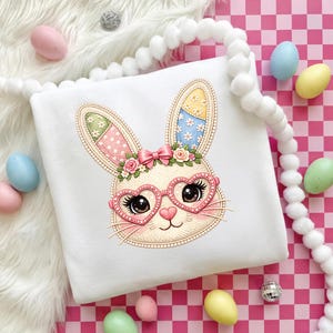Puede incluir: Sudadera blanca con temática de Pascua con un rostro de conejo bordado con gafas en forma de corazón, un lazo rosa y detalles florales. Las orejas del conejo tienen diseños de patchwork coloridos. Huevos de Pascua pastel y una guirnalda de pompones blancos rodean la sudadera.