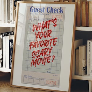May include: Wooden framed print with a guest check design. The print has the question "WHAT'S YOUR FAVORITE SCARY MOVIE?" in red. The print is displayed on a white shelf with books, a decorative basket, and a plant.