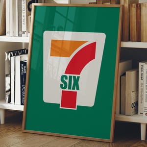 May include: A framed poster with a green background and a white, orange, and red logo. The word "SIX" is printed in green at the bottom of the logo. The poster is leaning against a white bookshelf.
