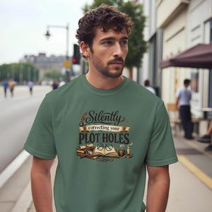 Peut inclure: T-shirt vert avec l'inscription "Silently correcting your PLOT HOLES". Le motif comprend un livre ouvert, des lunettes, des parchemins, une loupe et une bougie. Le t-shirt est porté par une personne.