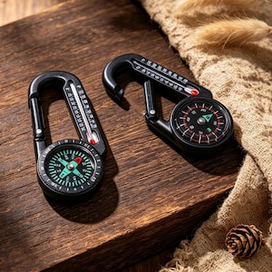 May include: Two black carabiner clips, each featuring an integrated circular compass with a green and black dial and a built-in thermometer along the side of the metal frame. The compasses display cardinal directions and degree markings clearly.