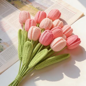 May include: A bunch of crochet tulips in shades of pink, with green stems and leaves. The handmade flowers are arranged on a newspaper background, creating a soft, textured appearance. The bouquet is tied together with green yarn.