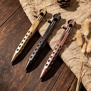 May include: Three bolt-action pens in brass, black, and copper finishes. Each pen features a perforated barrel design, a top-mounted bolt mechanism, and a pointed tip. The brass pen is engraved with the name Susan, and the copper pen with Rebecca.