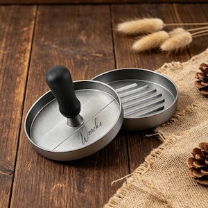 May include: A silver-toned metal burger press with a black handle and the name Wencke engraved on the top surface. The press consists of a circular base with a grooved interior and a matching lid with a central handle for shaping meat patties.