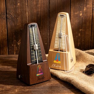 May include: Two wooden metronomes, one dark brown and one light wood, sit on a wooden surface. The dark brown metronome has the name "Jacob" and a colorful logo. The light wood metronome has the name "Christina" and a colorful logo. Sunglasses and a pinecone are also visible.