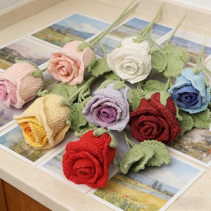 Op de afbeelding: Een verzameling handgemaakte gehaakte rozen in verschillende kleuren, waaronder roze, wit, geel, paars, rood en blauw. Elke roos heeft groene bladeren en stelen. De rozen zijn gerangschikt op een lichtgekleurd oppervlak.