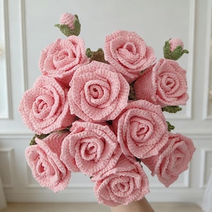 Könnte beinhalten: Ein Strauß handgefertigter, hellrosa gehäkelter Rosen. Die Rosen sind in voller Blüte, mit detaillierten Blütenblättern. Grüne Blätter und Knospen akzentuieren die Blumen. Der Strauß wird in einer Hand gehalten, mit weißem Hintergrund.