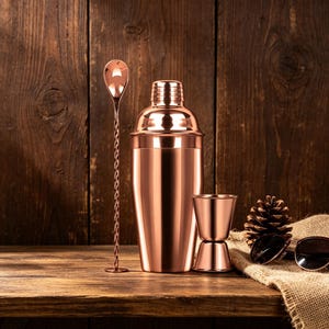May include: A copper-colored cocktail shaker set with a bar spoon and a double jigger. The set is displayed on a wooden surface with a wooden background. A pinecone and sunglasses are also present.