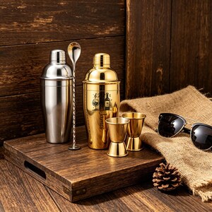 May include: A stainless steel cocktail shaker and a gold-colored cocktail shaker with a spoon, two jiggers, and sunglasses on a wooden tray. The gold shaker has a design and the name "James" on it. A pinecone sits on the tray.