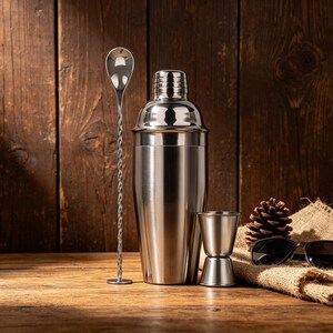 May include: A stainless steel cocktail shaker, a long bar spoon, and a double-sided jigger are displayed on a wooden surface. A pine cone and sunglasses rest on a burlap cloth in the background. The shaker is a classic Boston style.