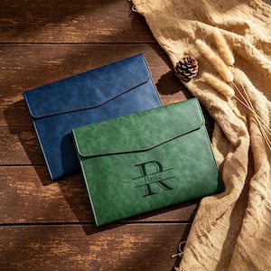 May include: Two leather document holders. One is a deep blue, the other is green with the name "Rose" and the letter "R" printed on the front. The holders are on a wooden surface with a burlap cloth and pinecone.