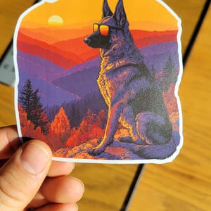 May include: A sticker featuring a German Shepherd wearing sunglasses, set against a sunset over a mountain range. The dog is depicted in a stylized, artistic manner, with vibrant colors of orange, red, and purple.
