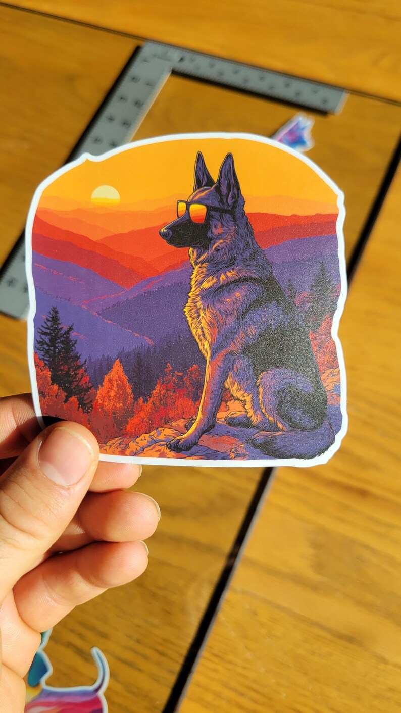 May include: A sticker featuring a German Shepherd wearing sunglasses, set against a sunset over a mountain range. The dog is depicted in a stylized, artistic manner, with vibrant colors of orange, red, and purple.