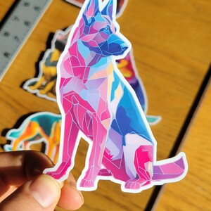 May include: A colorful sticker featuring a geometric design of a German Shepherd dog in shades of pink, blue, and purple. The sticker is held up by a hand, with other dog-themed stickers in the background.