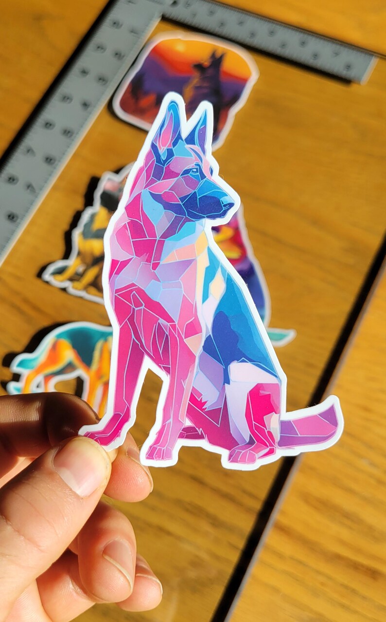 May include: A colorful sticker featuring a geometric design of a German Shepherd dog in shades of pink, blue, and purple. The sticker is held up by a hand, with other dog-themed stickers in the background.