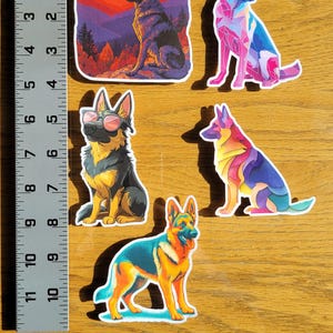 May include: A collection of colorful German Shepherd dog stickers. The stickers feature various artistic styles, including geometric and landscape designs. The stickers are on a wooden surface, with a ruler for scale.