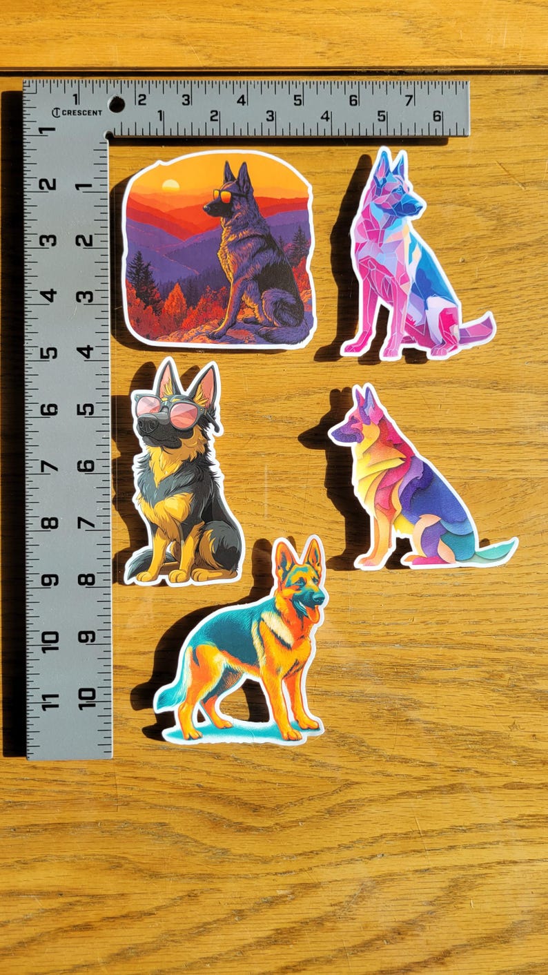 May include: A collection of colorful German Shepherd dog stickers. The stickers feature various artistic styles, including geometric and landscape designs. The stickers are on a wooden surface, with a ruler for scale.