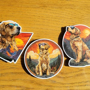 May include: Three stickers featuring a Golden Retriever wearing sunglasses. Each sticker has a different design, including a sunset, mountains, and trees. The dog is the central focus of each sticker.