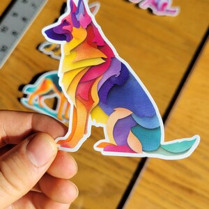 May include: A colorful sticker of a German Shepherd dog in a sitting position. The dog is depicted with layered, abstract shapes in shades of yellow, orange, pink, purple, and blue. Other dog-themed stickers are visible in the background.