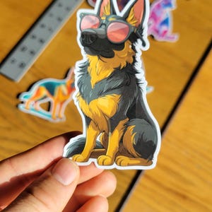 May include: A sticker featuring a cartoon German Shepherd wearing sunglasses with pink lenses. The dog is depicted in black and yellow, sitting upright. Other stickers are visible in the background.
