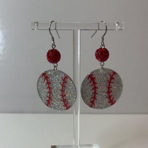 May include: A pair of drop earrings featuring circular, silver glitter baseball charms with red stitched detailing. Each earring is accented with a small, round, red rhinestone bead suspended from a silver-toned metal hook wire.