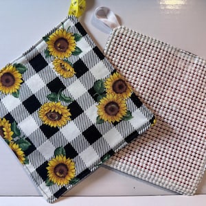 May include: Two square pot holders. One features a black and white buffalo check pattern with yellow sunflowers and green leaves, plus a yellow hanging loop. The other has a textured white and red grid pattern with a white hanging loop.