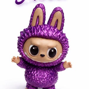 May include: A purple figurine of a creature wearing a textured, hooded outfit with bunny ears. The figurine has large eyes, a small nose, and a toothy grin. The name "Nova" is written above the figure in purple script.