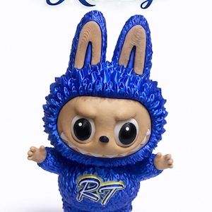 May include: A blue figurine of a creature wearing a bunny-eared hoodie. The figurine has large black eyes, a neutral expression, and small arms and feet. The text "Ritzy" is above the figurine, and the letters "RT" are on the chest.