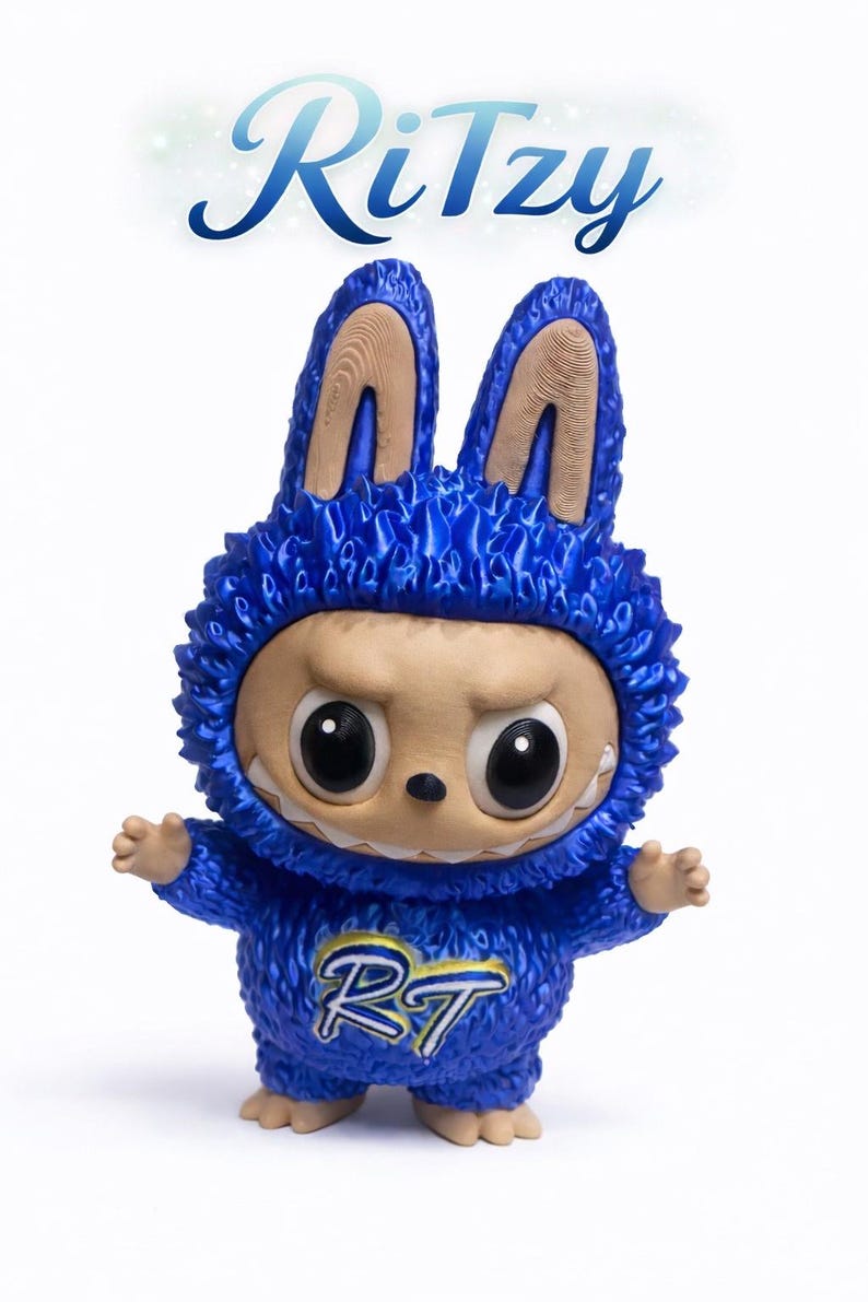 May include: A blue figurine of a creature wearing a bunny-eared hoodie. The figurine has large black eyes, a neutral expression, and small arms and feet. The text "Ritzy" is above the figurine, and the letters "RT" are on the chest.