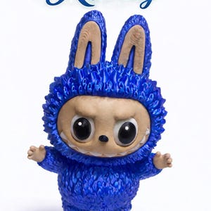 May include: A blue figurine of a creature wearing a bunny-eared hood with the word "Ritzy" above it. The figurine has large, expressive eyes, a neutral expression, and small arms and feet. The hood has a textured, furry appearance.