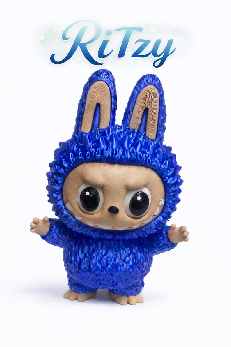 May include: A blue figurine of a creature wearing a bunny-eared hood with the word "Ritzy" above it. The figurine has large, expressive eyes, a neutral expression, and small arms and feet. The hood has a textured, furry appearance.