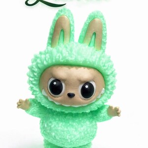 May include: A green plush figure in a bunny costume with the name "Luma" in green script above. The figure has large black eyes, a small nose, and a slightly menacing expression. The costume has a fluffy texture.