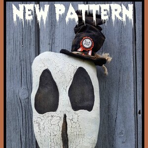 May include: A white ghost-shaped fabric doll with a black painted face and a black top hat with a red and white "Trick or Treat" badge. The doll is hanging on a weathered gray wooden wall.