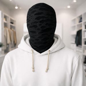 Puede incluir: Una máscara facial negra con un patrón desgastado se usa sobre la cabeza y el cuello. La persona lleva una sudadera con capucha blanca con cordones beige. El fondo es una tienda de ropa borrosa.