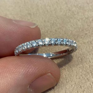 Puede incluir: Un anillo de banda de metal plateado con una fila continua de pequeñas piedras redondas transparentes talladas a lo largo de la mitad superior de la banda. El anillo tiene un acabado pulido y refleja la luz en las piedras y el metal.