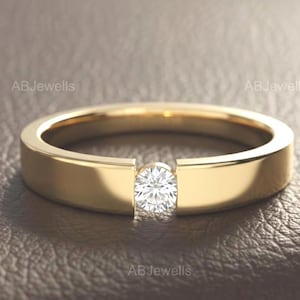Könnte beinhalten: Ein goldener Bandring mit einem einzelnen runden, im Brillantschliff geschliffenen weißen Diamanten in einer Spannfassung. Der Ring hat eine glatte, polierte Oberfläche und ein flaches Profil. Der Text ABJewells ist links und rechts zu sehen.