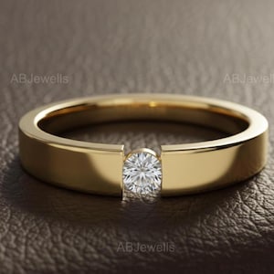 Könnte beinhalten: Ein goldener Ring mit einem einzelnen, runden, klaren Edelstein in der Mitte. Das Ringband ist flach und poliert. Der Ring wird auf einer braunen Oberfläche präsentiert. Der Text "ABJewells" ist sichtbar.