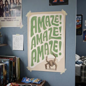 Puede incluir: Un p&oacute;ster de pared rectangular que presenta la palabra AMAZE! repetida tres veces en letras grandes, onduladas y verdes sobre un fondo crema. Debajo del texto, una peque&ntilde;a criatura de color canela con varias patas.