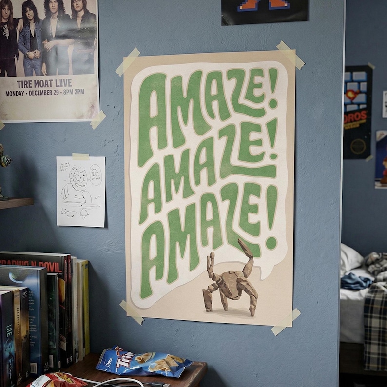Puede incluir: Un p&oacute;ster de pared rectangular que presenta la palabra AMAZE! repetida tres veces en letras grandes, onduladas y verdes sobre un fondo crema. Debajo del texto, una peque&ntilde;a criatura de color canela con varias patas.