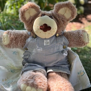 May include: A brown teddy bear wearing blue and white striped overalls with gold-coloured buttons. The bear has a light brown snout and large, fluffy ears. The bear is sitting on a white patterned blanket.