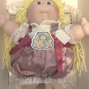 May include: A Cabbage Patch Kids doll with yellow yarn hair, green eyes, and a soft fabric body. The doll wears a mauve satin romper with a floral pattern, white lace trim, and a white collar. A Cabbage Patch Kids tag is attached to the chest.
