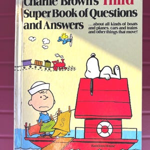 May include: A vintage copy of "Charlie Brown's Third Super Book of Questions and Answers." The cover features Charlie Brown, Snoopy, and a red doghouse on a boat. The book's title is in large black letters with "Third" in red.